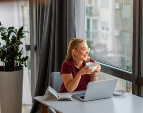 Image of a woman at home with a laptop, book, and coffee, looking relaxed.