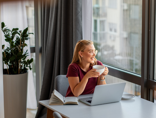 Image of a woman at home with a laptop, book, and coffee, looking relaxed.