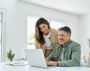 Image of a middle-aged man working on a laptop next to a woman, representing will preparation.