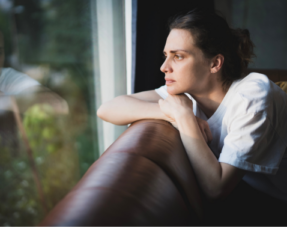A woman sitting on a couch, looking thoughtfully out a window.